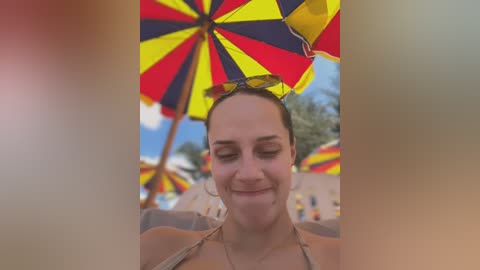 Media: Video of a smiling woman with brown hair under a large, colorful beach umbrella. She is wearing a beige bikini, with the beach in the background featuring palm trees and more umbrellas.