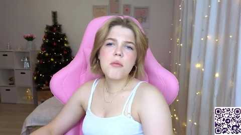 Video of a young woman with shoulder-length blonde hair, fair skin, wearing a white tank top, sitting in a pink gaming chair. Background features a decorated Christmas tree and string lights, a white shelving unit, and a QR code watermark.