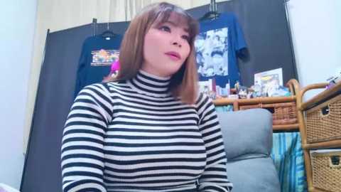 Media: Video of a woman with shoulder-length brown hair, wearing a black-and-white striped turtleneck, sitting in a room with blue and black shirts hanging, wicker furniture, and a beige wall.