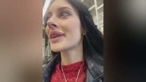 Media: Video of a smiling, light-skinned woman with long black hair, wearing a red lace top and black leather jacket, with a blurred, dimly lit background.