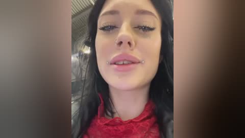 Media: Video of a young woman with long black hair, fair skin, and a silver nose ring, wearing a red lace top, captured from an up-close angle indoors.