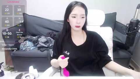 Media: Video of an East Asian woman with long black hair, wearing a black shirt, sitting at a desk. She holds a pink vibrator. Background includes a gray couch, a TV, and a cluttered desk.