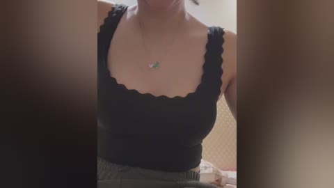 Media: Video of a woman in a black, ruffled sleeveless top with a small green pendant necklace, partially obscured by out-of-focus objects on the sides.