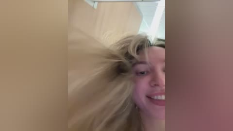 Media: Video of a young woman with light skin and blonde hair, smiling, partially obscured by a brown curtain. Background shows a bathroom with white tiles and a ceiling light.