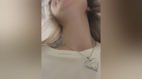 Media: Video of a woman with light skin and blonde hair, wearing a white shirt with a heart pendant necklace. She's smiling, her head tilted back, with blurred background.