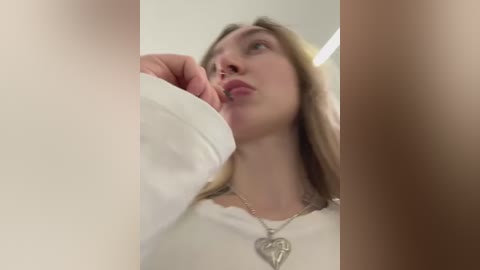 Media: A video of a young woman with light skin and long blonde hair, wearing a white shirt and a heart necklace, looking up thoughtfully. The image is cropped closely, focusing on her face and upper body.