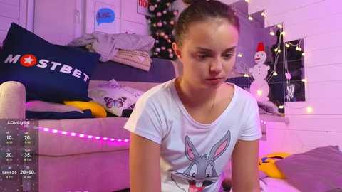 Media: Video of a young girl with light skin, brown hair in a bun, wearing a white t-shirt with a cartoon bunny, sitting indoors next to a sofa with a blue pillow. Christmas decorations and string lights in the background.