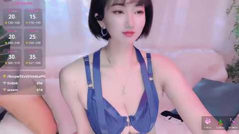 Media: Video of a young Asian woman with short black hair, wearing a blue halter top, sitting on a white couch. Background features a potted plant and a blue pillow.