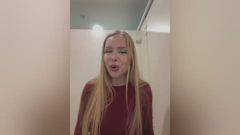 Video of a young Caucasian woman with long, straight blonde hair, wearing a maroon sweater, standing in a narrow, beige-tiled bathroom with a metal pipe and overhead light.