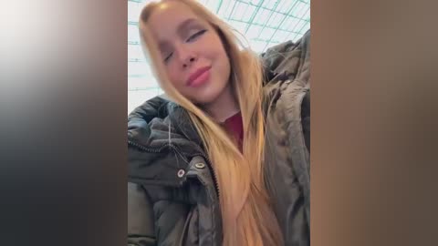 Media: Video of a blonde woman with fair skin, wearing a gray puffy jacket, eyes closed, smiling, in a room with a grid-tiled ceiling.