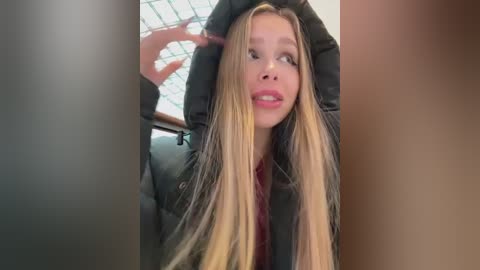 A video of a blonde woman with long hair, wearing a black coat and red shirt, sitting in a car with a grid-tiled ceiling, slightly blurry background.