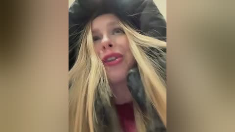 Media: Video of a blonde woman with fair skin, wearing a black puffy jacket and red lipstick, smiling while playfully tugging her hair.