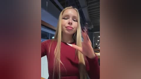 Video of a blonde, fair-skinned young woman with long straight hair, wearing a maroon long-sleeved shirt, standing in a modern indoor setting with exposed ceiling beams and glass windows, holding a black hairbrush.