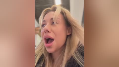 Media: Video of a blonde woman with tousled hair, open mouth in a surprised or shocked expression, wearing a dark jacket. Background is blurred, showing a bright, indoor setting.