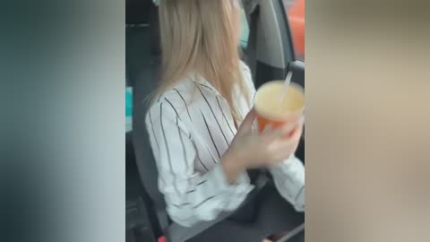 Media: Video of a woman with long blonde hair, wearing a white and black striped blouse, holding a drink, seated in a car.