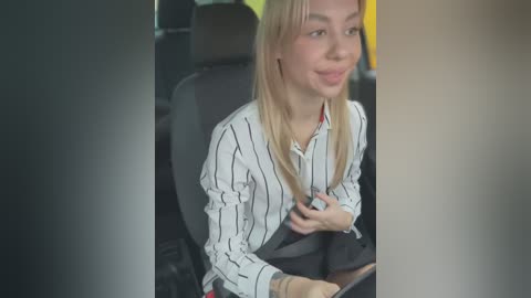 Media: Video of a blonde Caucasian woman in a white striped blouse, wearing a black seatbelt, seated in a car with dark upholstery, looking relaxed.