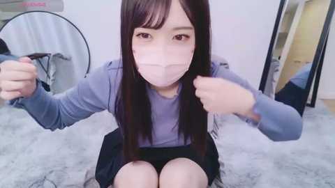 Media: Video of an Asian woman with long black hair, wearing a light purple hoodie and mask, sitting on a fluffy white rug, fists clenched, in a room with a mirror and a white circular light fixture.