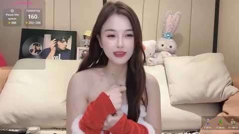 Media: A young Asian woman with long dark hair, wearing a red Santa costume, sits on a beige sofa with a stuffed bunny, in a cozy, softly lit living room.