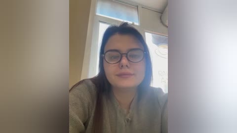 Media: Video of a young woman with long brown hair, wearing glasses, light beige top, indoors near a window with bright light.