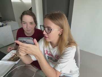 Media: Video of two Caucasian teenage girls, one with glasses, sitting at a table. They are engrossed in a smartphone, wearing casual t-shirts. Background includes a white door and a cluttered white shelf.