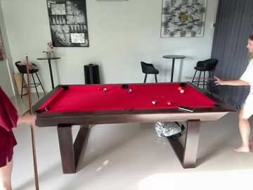 Media: Video of a modern billiards room with a vibrant red pool table, three black stools, black-and-white artwork, and a partially visible person in a white shirt.