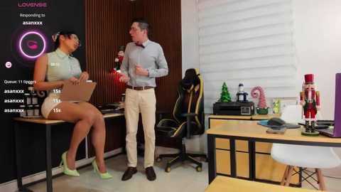 Media: Video of a modern office scene: a woman in a tight green top and short skirt presents, while a man in a blue shirt and beige pants listens attentively. Background includes a black wall, white blinds, and office decor.