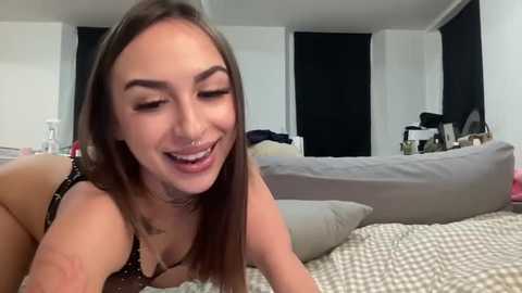 Media: A video of a young woman with straight, shoulder-length brown hair, wearing a black lace bra, lying on a bed with a checkered duvet. She has a light complexion and a smiling expression.