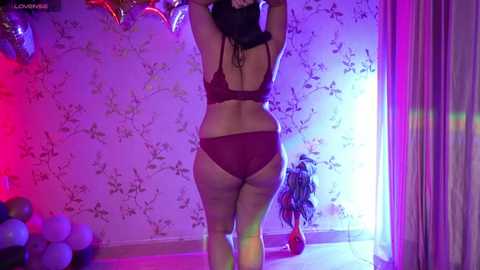 Media: Video of a curvy woman with medium skin tone, wearing maroon lingerie, standing with arms raised, in a dimly lit room with purple and pink lights, floral wallpaper, and a plant.