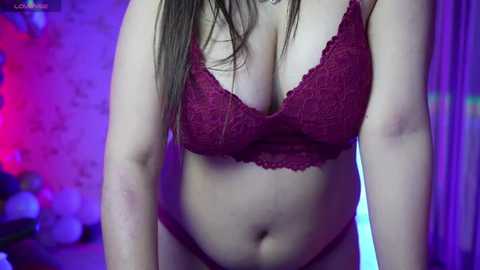 Media: Video of a woman in a red lace bra, revealing medium-sized breasts, with her long brown hair partially visible. Background features purple and pink lighting, blurred objects, and a brick wall.