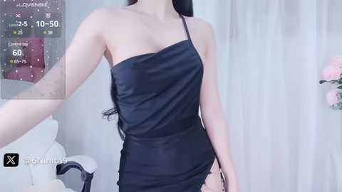 Media: A video of an East Asian woman in a black, one-shoulder mini dress, standing in a soft-lit, minimalist room with a floral backdrop.