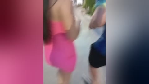 Media: A blurred video of a woman in a tight, bright pink dress, walking away from the camera. The background features a blurred street scene with green foliage and a person in a blue shirt.