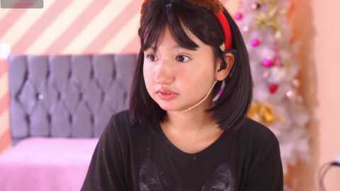 Media: Video of an Asian woman with straight black hair, wearing a black t-shirt, in a cozy, pink-themed room with a gray tufted headboard, Christmas decorations, and a pink bed.