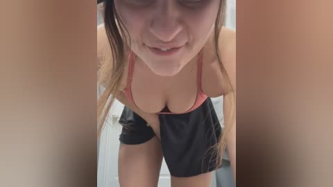 Media: Video of a young, fair-skinned woman with long brown hair, wearing a red bra and black shorts, leaning forward with a playful expression, taken from a low angle.