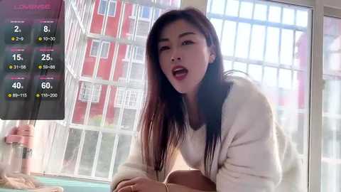 Video of an East Asian woman with long black hair, wearing a white sweater, looking surprised in a modern apartment with large windows and red brick walls.