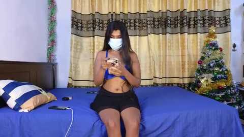 Media: Video of a young woman with long dark hair, wearing a blue crop top, black shorts, and a face mask, sitting on a bed, holding a phone, in a room with a Christmas tree and yellow curtains.