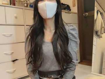 Media: Video of a woman with long, wavy black hair, wearing a blue face mask, gray top, and black belt, standing in a cluttered, white-cabinet-filled room.