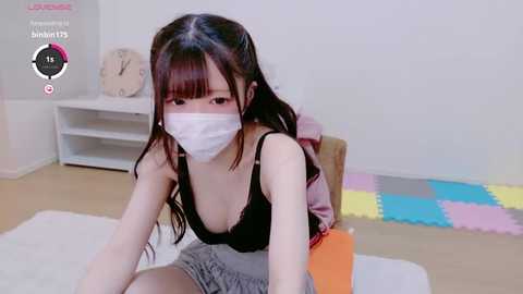 Media: A video of an Asian woman with long brown hair, wearing a face mask and black tank top, kneeling on a white rug in a minimalist room with colorful play mats and a clock.