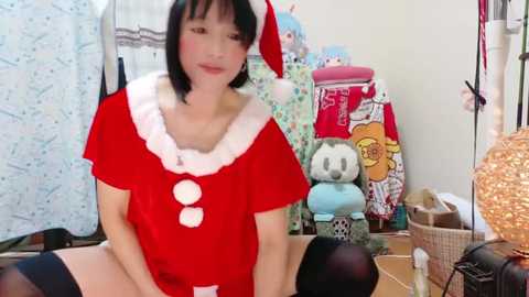 Media: A video of an Asian woman with black bobbed hair, wearing a red Santa costume, sitting in a cluttered room with stuffed animals and Christmas decor.