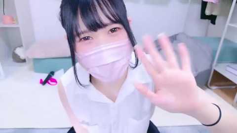 Media: Video of an Asian woman with pale skin, dark hair, and a white face mask, waving from a distance, in a minimalist room with pastel hues.