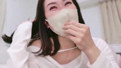 Media: Video of an East Asian woman with long black hair, wearing a white mask, a white top, and a necklace, indoors, smiling with closed eyes, blurred background.