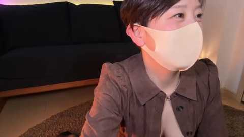 Media: Video of an East Asian woman with short black hair, wearing a beige face mask, grey jacket, and seated indoors, with a black sofa and woven rug in the background.