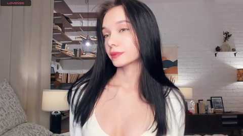 Video of a young woman with long black hair, fair skin, and red lipstick, wearing a white blouse, in a cozy living room with white brick walls, wooden shelves, and a lamp.