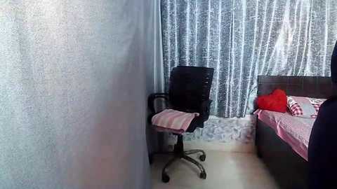 Media: Video of a small, sparsely furnished room with a black office chair and a bed with pink and red checkered bedding. Gray textured walls, white curtains with floral patterns, and a carpeted floor.