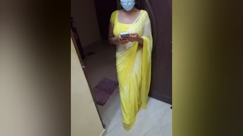 Media: Video of a woman in a yellow sari, holding a smartphone, wearing a face mask, standing in a dimly lit corridor with beige walls and a purple rug.