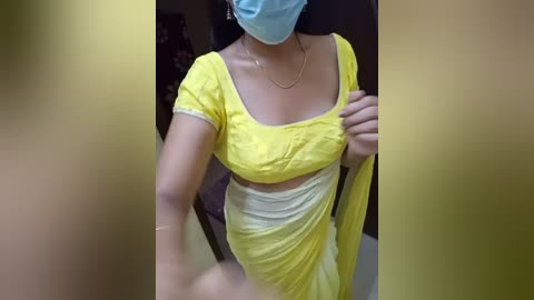 Media: Video of a woman in a yellow saree and blue mask, with a blurred background, showcasing a casual indoor setting.