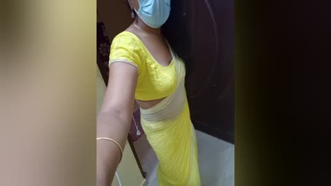 Media: A video of a person in a bright yellow dress with a large cutout, wearing a blue face mask and a bracelet, standing against a dark background.