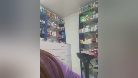 A video of a cluttered, well-lit room with a purple bed, white dresser, and shelves packed with colorful medicines.