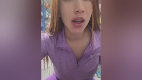 Media: Video of a young woman with fair skin, straight auburn hair, and light makeup, wearing a purple zip-up jacket. She's indoors, with blurred shelves of various items in the background. Her lips are slightly parted, and she appears to be looking down.