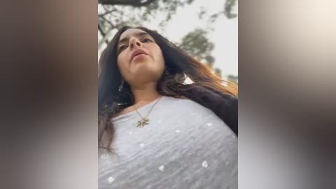 Media: Video of a young woman with medium-brown skin, long dark hair, wearing a white shirt with small water droplets, against a blurred green background.