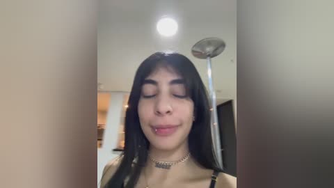 Media: Video of a young woman with long black hair, closed eyes, and a slight smile, wearing a choker necklace, standing in a modern kitchen with stainless steel appliances and a ceiling light.